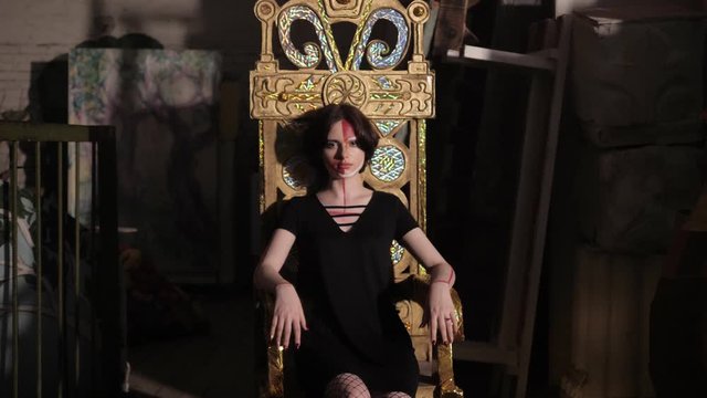 attractive young woman with red paint on face sits on decorative throne in room full of old things slow motion