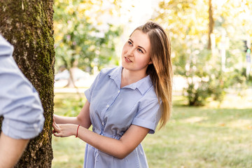 Fototapeta premium Young caucasian blond woman in a blue stripped summer dress playing hide and seek with her boyfriend in the park outdoors in a sunny day