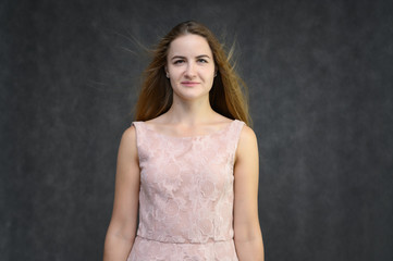 Obraz premium Studio portrait on the waist of a pretty student girl, a young brunette woman with long beautiful hair in a pink dress on a gray background. Smiling, talking, showing emotions