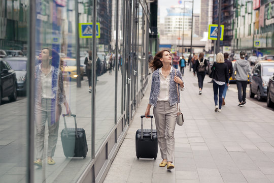 attractive elegant young woman in business clothes walks along the street along a glass building and rolls a small suitcase on wheels