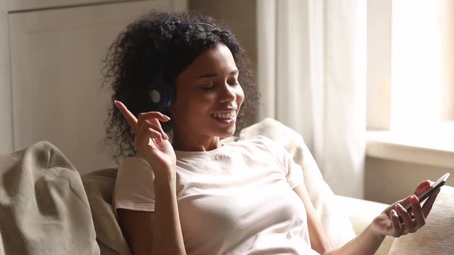 African woman leaned on couch listening music using smartphone headphones