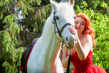 Portrait of a beautiful girl with red hair in a red dressy dress near with a white horse on a green...