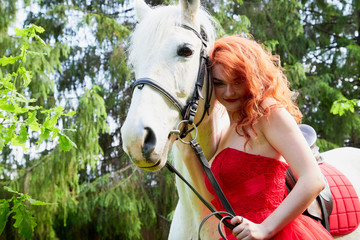 Portrait of a beautiful girl with red hair in a red dressy dress near with a white horse on a green...
