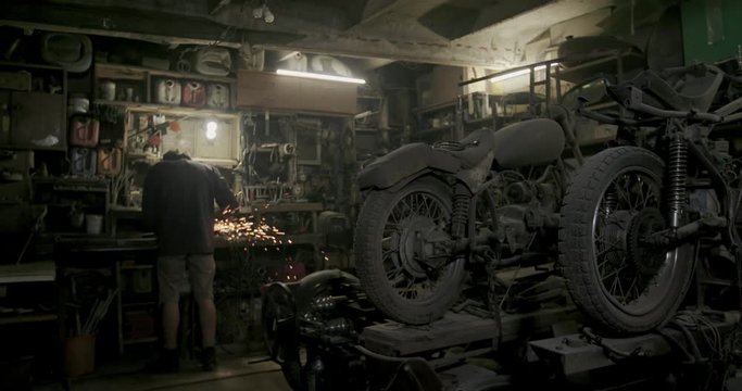 Metalworker working on a grinder with flying fiery sparks in a workshop with rows of stored vintage motorbikes in dim light