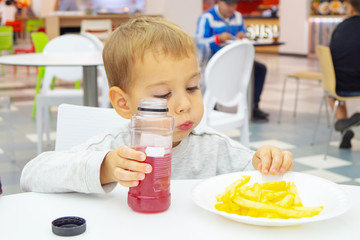 little child eats french fries and drinks juice, sitting at the table on the food court of the mall. Unhealthy food.