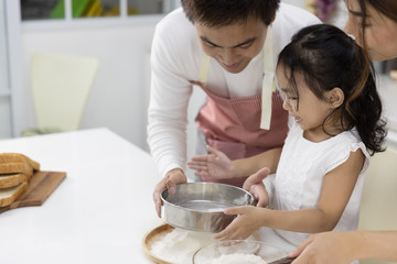 Happy Asian family Father, Mother and Daughter are sieving flour preparing the dough, bake cookies in the kitchen. family cooking food Concept.