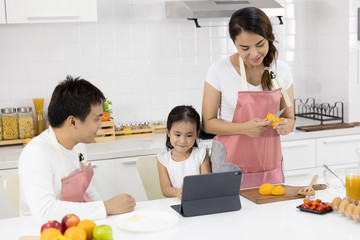 Obraz na płótnie Canvas Happy Asian family make a cooking. Father, Mother and Daughter are preparing fruit orange and see Digital tablet in the kitchen at home. Healthy food concept and happy holidays
