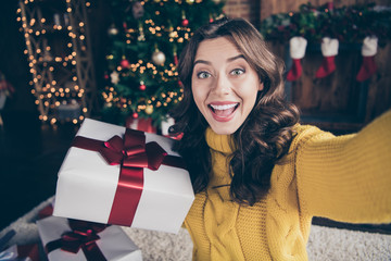 Self portrait of cheerful rejoicing girl shooting her gift box with christmas tree behind wearing yellow sweater