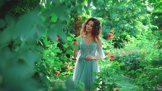 A woman in a luxurious evening dress of turquoise color walks in the garden among blooming roses. Princess with stylish styling on her loose hair. Fashionable image of a graduate girl at a prom party
