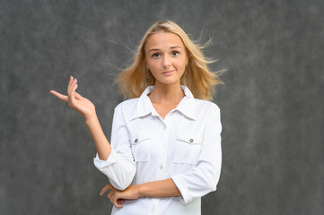 Studio portrait of a pretty blonde student girl, young woman in a white shirt on a gray background. Talking, showing emotions.
