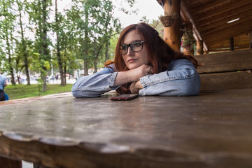Very pretty redhair girl in glasses and denim jacket sitting in cave and put her head on arms crossed on the table. Smartphone near her face on the table