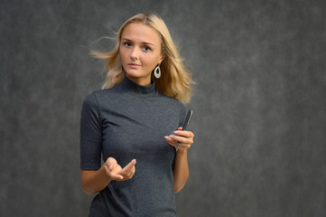 Studio portrait of a pretty blonde student girl, young woman in a gray sweater with a pen in her hands on a gray background. Talking, showing emotions.