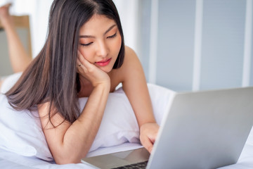 Fototapeta premium Pretty attractive sensuality young Asian woman using laptop, laying on the white bed at bedroom in the morning