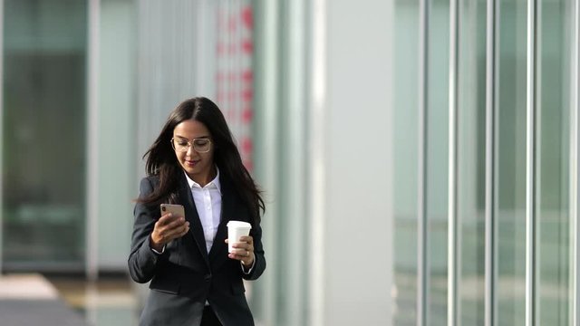 Smiling Asian woman using smartphone during stroll. Cheerful business lady walking with paper cup and modern phone. Technology concept