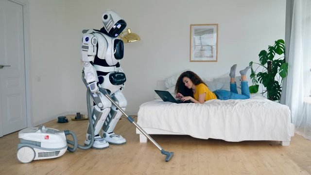 Girl works with a laptop while a cyborg vacuum cleans. Robot and human cooperation.