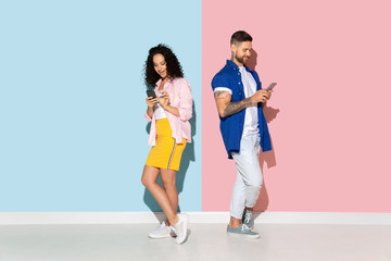Young emotional caucasian couple in bright casual clothes posing on pink and blue background. Concept of human emotions, facial expession, relations, ad. Woman and man using smartphone, look busy.