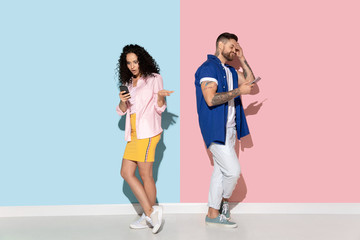 Fototapeta na wymiar Young emotional caucasian couple in bright casual clothes posing on pink and blue background. Concept of human emotions, facial expession, relations, ad. Woman and man using smartphone, look upset.