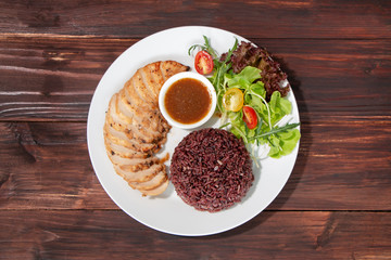 Top view of roasted chicken breast fillet steak with brown rice/coarse rice, vegetables salad and spicy sauce in white plate on wooden table. Healty food menu, eating right concept.