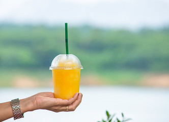 Hand holding a glass of cold Orange juice smoothie Background blurry views tree and water.