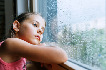 Little sad girl pensive looking through the window glass with a lot of raindrops. Sadness childhood...