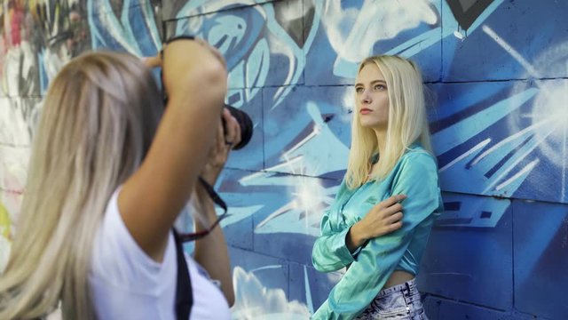 Blond girl poses for a girl photographer at the wall with graphitti in the summer in the city. Beautiful young girl model posing for a girl photographer. Graffiti wall. 4k