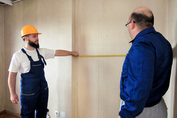 Engineer, contractor and project manager with his colleague, construction worker, handyman and builder is measuring wall room of apartment for renovation using measure tape at construction site.