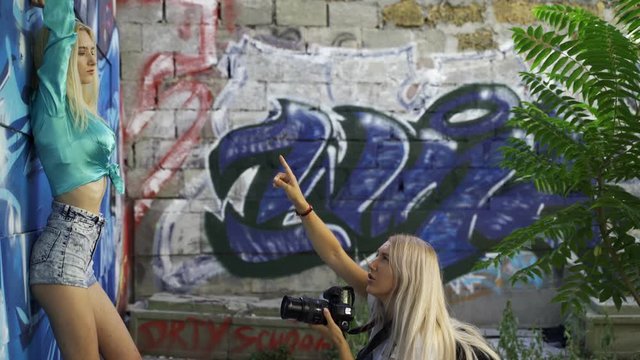 Blond girl poses for a girl photographer at the wall with graphitti in the summer in the city. Beautiful young girl model posing for a girl photographer. Graffiti wall. 4k