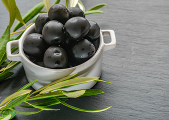 Close up of bowl filled with freshly harvested whole black olives. Branches with leaves and olives on a grey stone background with copy space for your text. Natural background. ECO products.