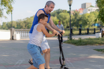 Young father teaching son to ride scooter in summer park.
