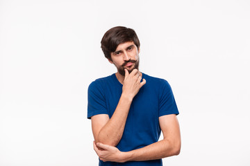 Thoughtful handsome brunet man in a blue shirt holding arms folded over his chest and keeping one hand close to face standing isolated over white background.