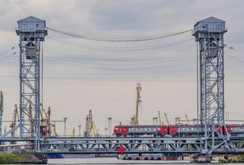 Passenger train crosses the bridge against the background of port cranes