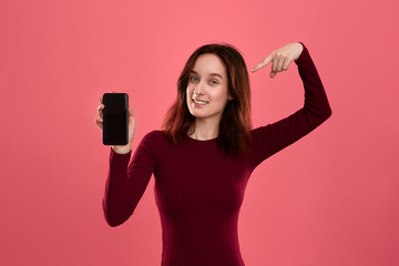 Excited pretty brunette girl pointing at the screen of a mobile phone with a finger while standing on a dark pink background looking at the camera.