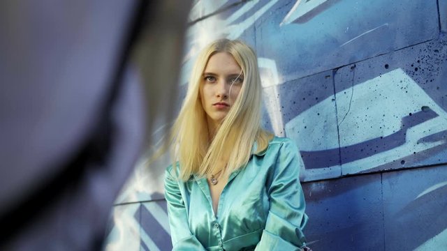 Blond girl poses for a girl photographer at the wall with graphitti in the summer in the city. Beautiful young girl model posing for a girl photographer. Graffiti wall. 4k