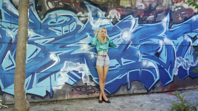 Blond girl poses for a girl photographer at the wall with graphitti in the summer in the city. Beautiful young girl model posing for a girl photographer. Graffiti wall. 4k
