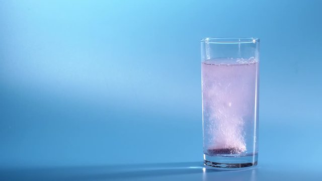 Effervescent Tablet Falling In Water. Pill Falls And Dissolves With Bubbles. Effervescent Tablet In Glass Of Water On Blue Background. Shot Of Effervescent Pill Dissolving In A Glass Of Water.