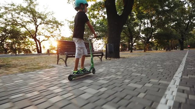 camera follows young skater riding scooter along green park paved road with brown wooden benches slow motion