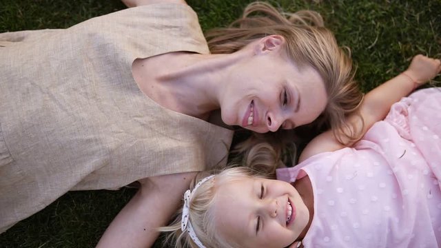 Young graceful blonde woman with her little blondie daughter, relaxing on grass in park. The mom gives a kiss to her baby and looking on her with joy and love. High angle view. Slow motion