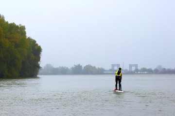 Fototapeta na wymiar Athlete paddling SUP (Stand up paddle board) at Danube river at cold weather against overcast sky. Concept of water tourism, water sport, healthy lifestyle and recreation