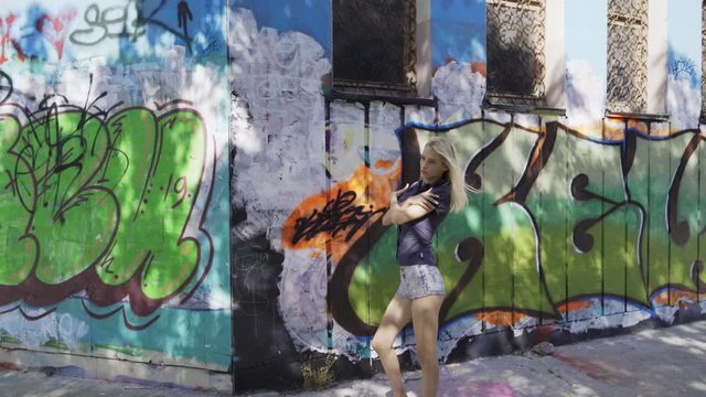 Blond girl poses for a girl photographer at the wall with graphitti in the summer in the city. Beautiful young girl model posing for a girl photographer. Graffiti wall. 4k