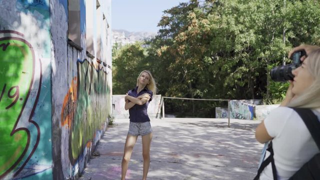Blond girl poses for a girl photographer at the wall with graphitti in the summer in the city. Beautiful young girl model posing for a girl photographer. Graffiti wall. 4k