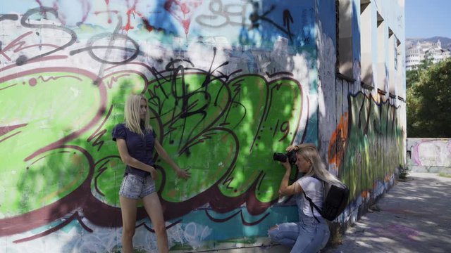 Blond girl poses for a girl photographer at the wall with graphitti in the summer in the city. Beautiful young girl model posing for a girl photographer. Graffiti wall. 4k