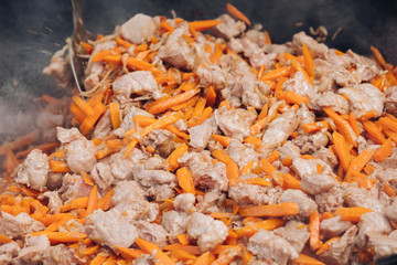 Process of cooking of delicious pilaf. View from above of tasty pieces of meat and healthy carrots roasting in cauldron during picnic in garden. Concept of cuisine and food.