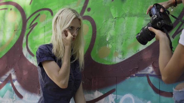 Blond girl poses for a girl photographer at the wall with graphitti in the summer in the city. Beautiful young girl model posing for a girl photographer. Graffiti wall. 4k