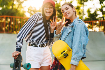 Image of two cheerful girls gesturing peace sign while poising with skateboards in skate park