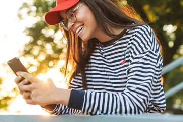 Image of laughing caucasian girl smiling and using cellphone while bending on railing in green park