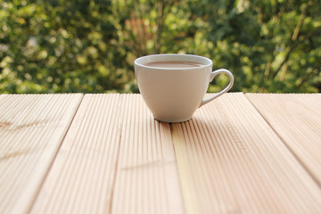 white cup with coffee on a light wooden table against a green summer garden, close-up, copy space
