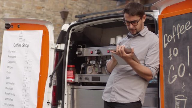 Thigh-up arc shot of frowning Caucasian male barista in glasses leaning against rear door of his mobile coffee van parked in city center street, using tablet computer, and waiting for customers