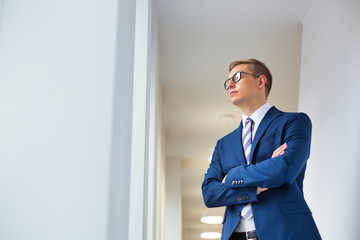 Fototapeta premium Young thoughtful businessman standing with arms crossed looking away in office corridor
