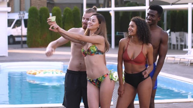 Smiling attractive multinational young people in swimsuits posing for selfie shot on background of swimming pool in luxury spa hotel. Joyful diverse friends having fun taking pictures by outdoor pool
