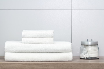 Four white towels of different sizes neatly folded lie on a wooden shelf and next to it is a can with cotton pads and ear sticks against a ceramic tile.